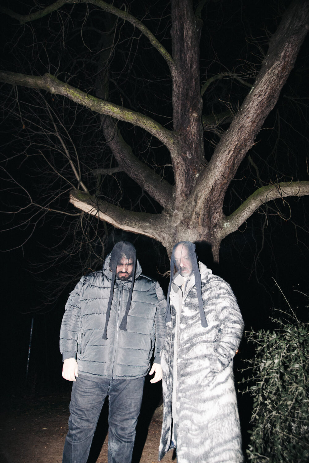 Zwei personen stehen nachts in einem Park. Sie tragen Strumpfhosen über dem Kopf, um ihre Gesichter zu verzerren.