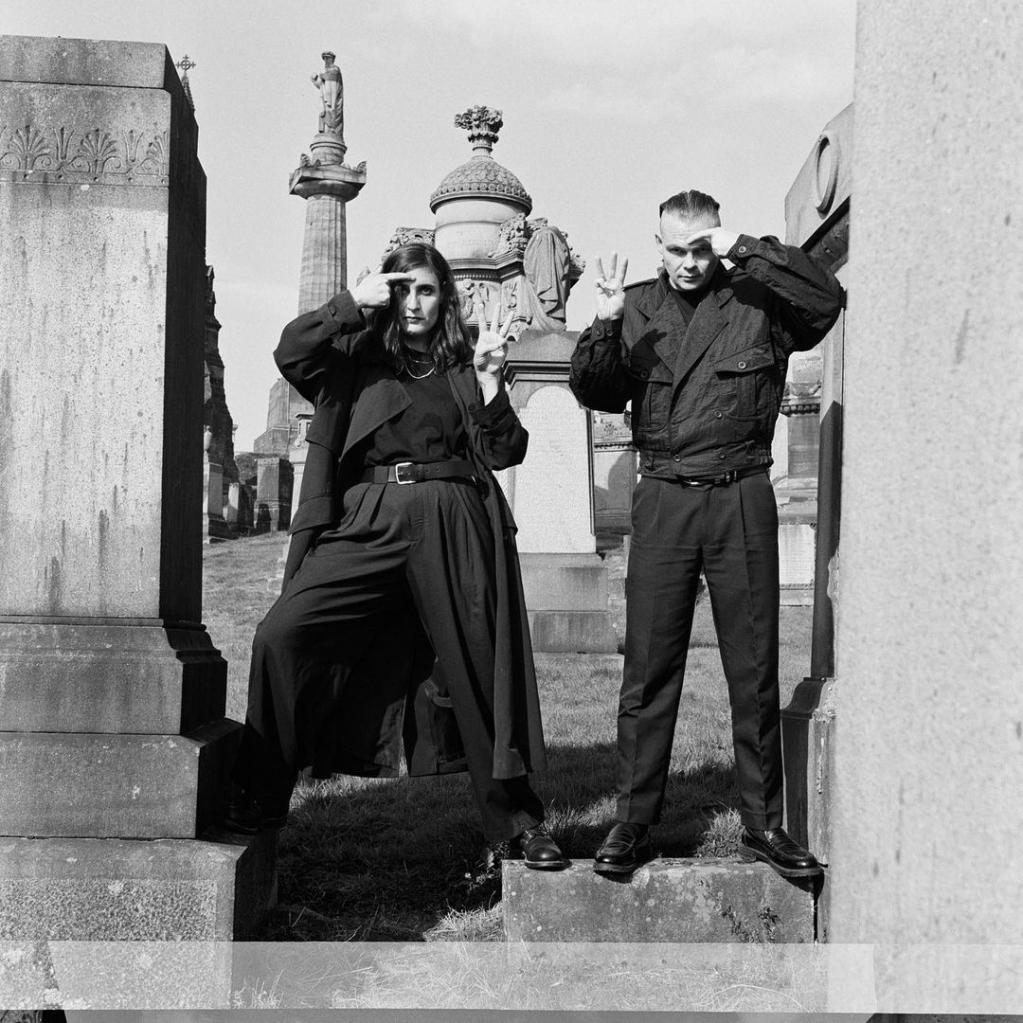Ein schwarz-weiß Bild von zwei Menschen, die auf einem alten Monument stehen un ihre Augen mit einer Hand vor der Sonne schützen.