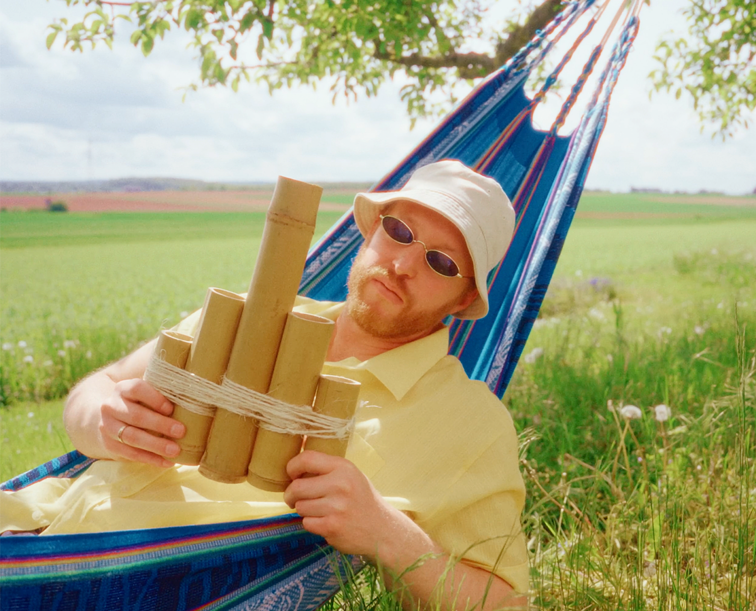 Ein Mann mit Hut und Sonnenbrille liegt in einer Hängematte. Er hält in beiden Händen eine Panflöte, die an einen ausgestreckten Mittelfinger erinnert.