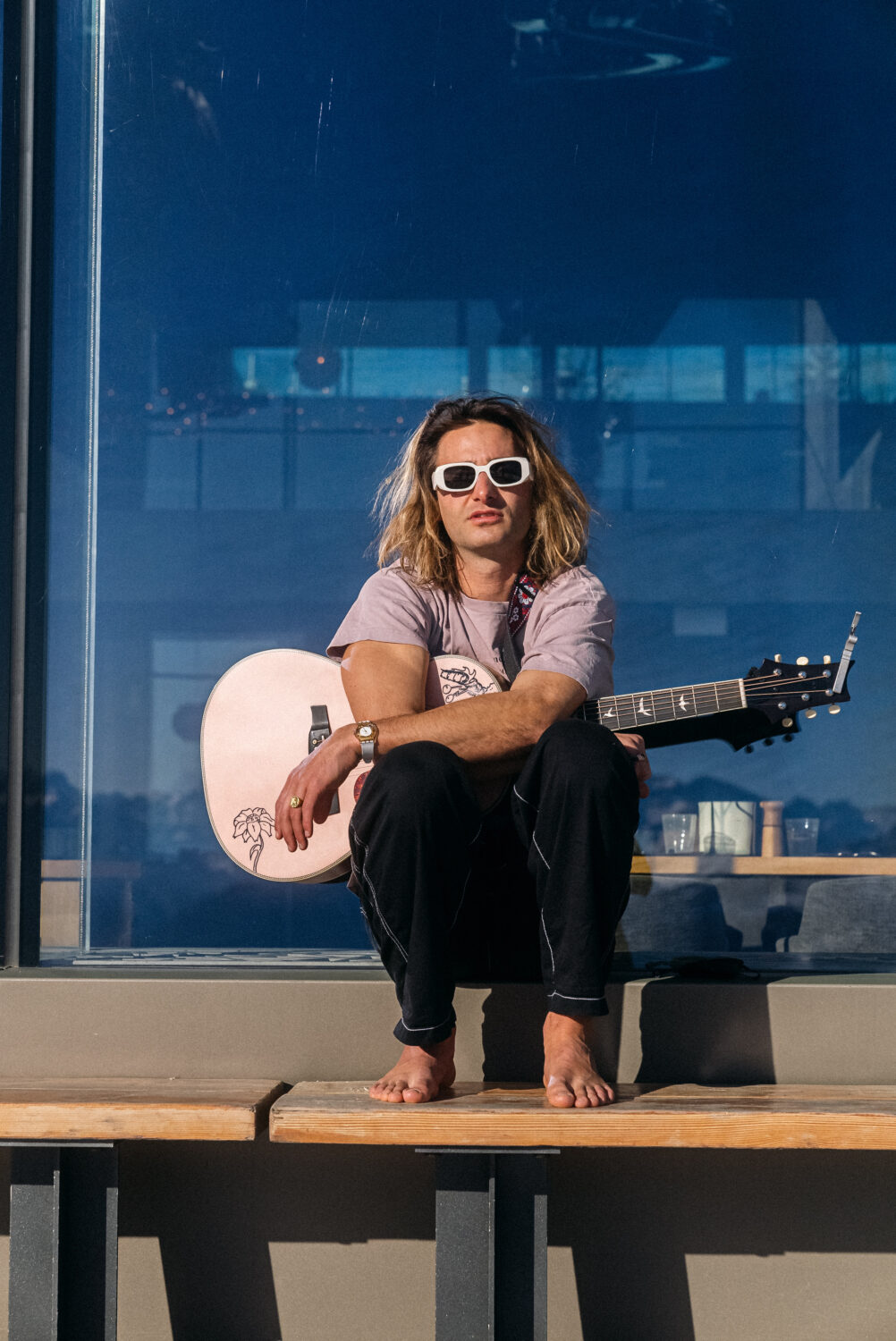 Ein Mann mit langen Haaren und einer gitarre auf dem Schoß sitzt in der Sonne.