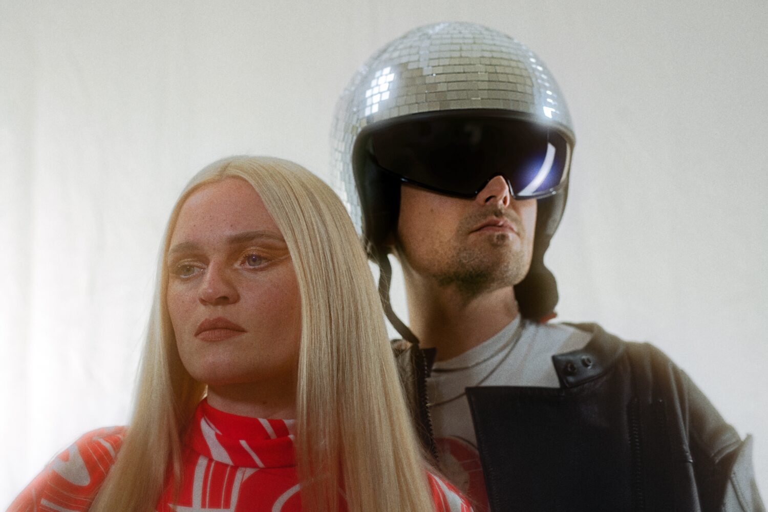 Ein Bild von einer blonden Frau im roten Kleid und einem Mann mit Sonnenbrille und Helm. Der Helm ist eine Discokugel.