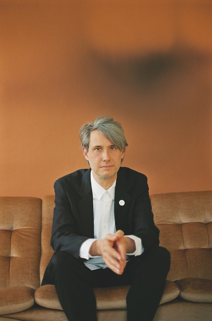 Ein Mann im Anzug sitzt auf einer braunen Ledercouch vor einem organgen Hintergrund.