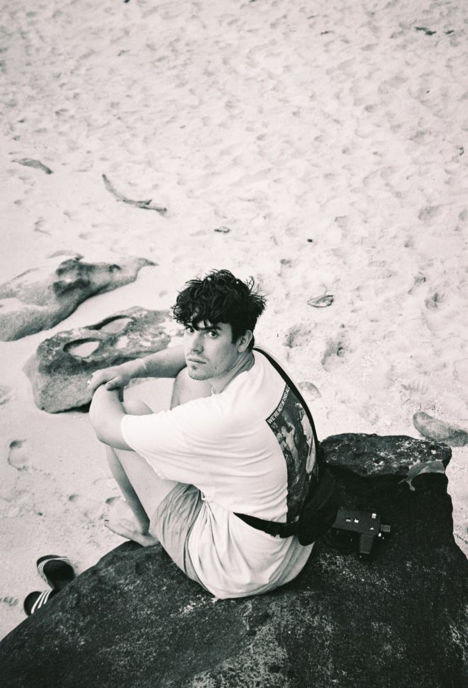Ein schwarz-weiß Bild von einem jungen brünetten Mann, der auf einem Fels am Strand sitzt.
