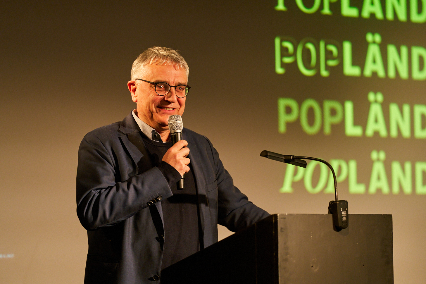 Das Bild zeigt den Staatssekretär Arne Braun. Er steht in einem schwarzen Anzug mit schwarzer brille hinter einem Rednerpult und spricht in ein Mikro. Hinter ihm ist eine Projektion des Popländ-Logos zu sehen.