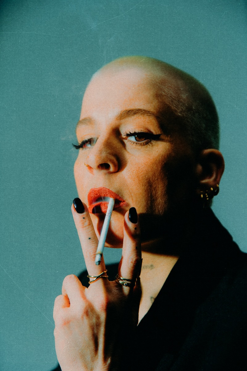 Das Bild zeigt Kiki. Die Musikerin trägt abrasiertes Haar und lange künstliche Wimpern. Sie blickt direkt in die Kamera und zieht dabei lasziv an einer Zigarette, die ihr aus dem linken Mundwinkel hängt.