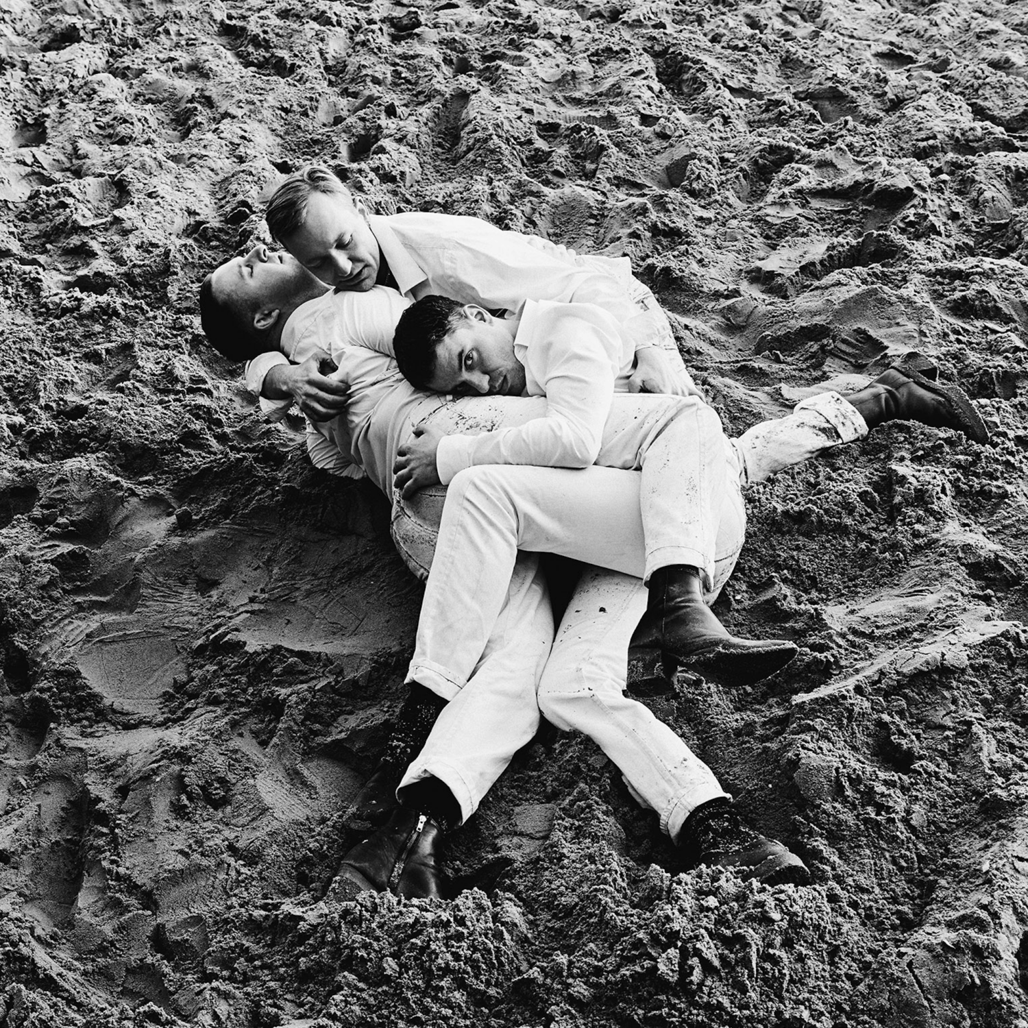 Das Bild zeigt das Trio die Selektion. Drei Männer in weißen Anzügen schmiegen sich im Sand liegend aneinander.