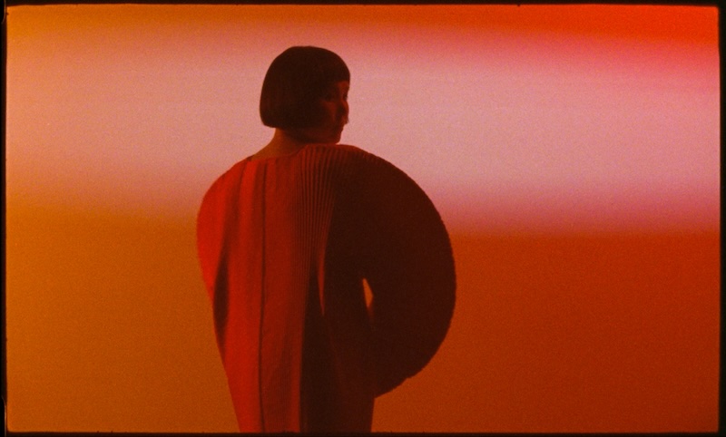 Das Bild zeigt die Musikerin Enji. Sie dreht der Kamera den Rücken zu trägt ein übergroßes rotes Kleid und blickt dabei über ihre Schulter.