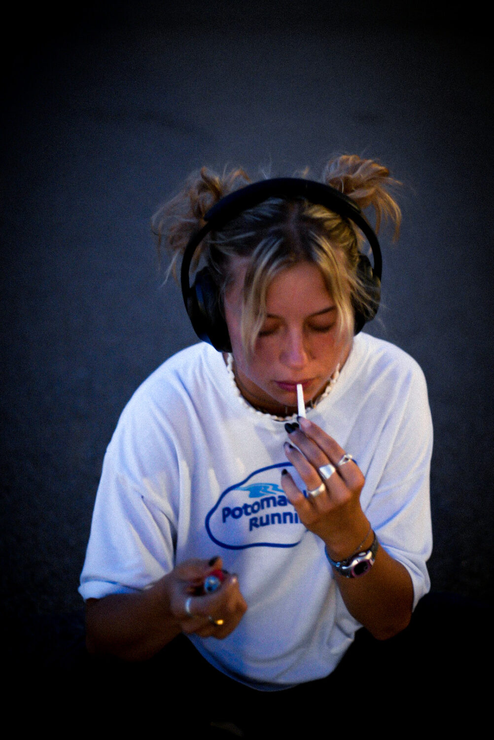 Das Foto zeigt die Musikerin Mara. Sie trägt zwei blonde Zöpfe, ein weißes Shirt, viele Ringe an den Fingern und Kopfhörer. Sie zündet sich gerade eine Zigarette an, ihr blick ist auf ihre Hand mit einem feuerzeug gerichtet.