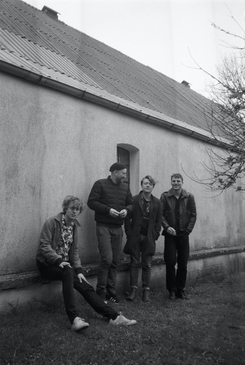 Das Bild zeigt die Band Trümmer. Das Foto ist schwarz weiß und zweigt die vier Bandmitglieder, wie sie entspannt nebeneinander vor einer Hauswand stehen.