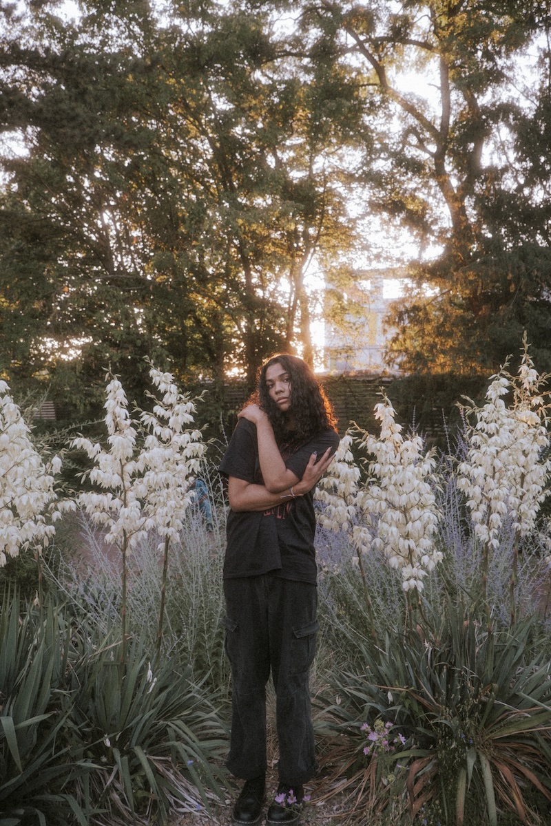 Das Bild zeigt die Musikerin Absteige. Sie steht zwischen schulterhohen weißen Blumen und trägt schwarze Kleidung und langes dunkles Haar. Sie hat ihre Arme um ihre Schultern geschlungen und blickt in die Kamera.