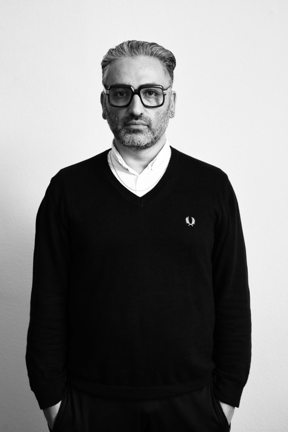 Ein Mann mit Brille und Pullover steht vor einer weißen Wand.