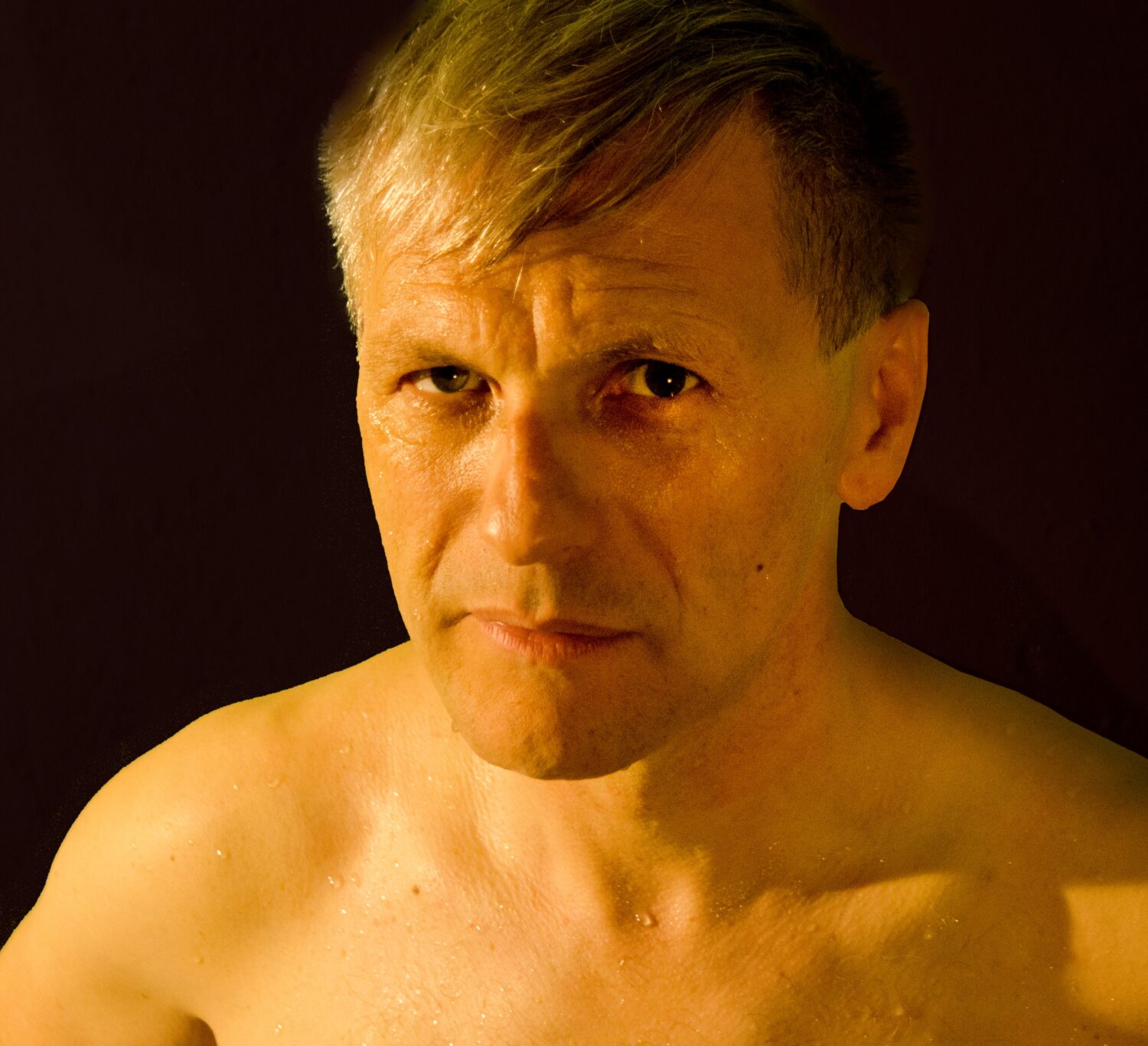 Ein muskulöser Mann ohne Hemd steht vor einer schwarzen Wand mit einem entspannten Gesichtsausdruck.